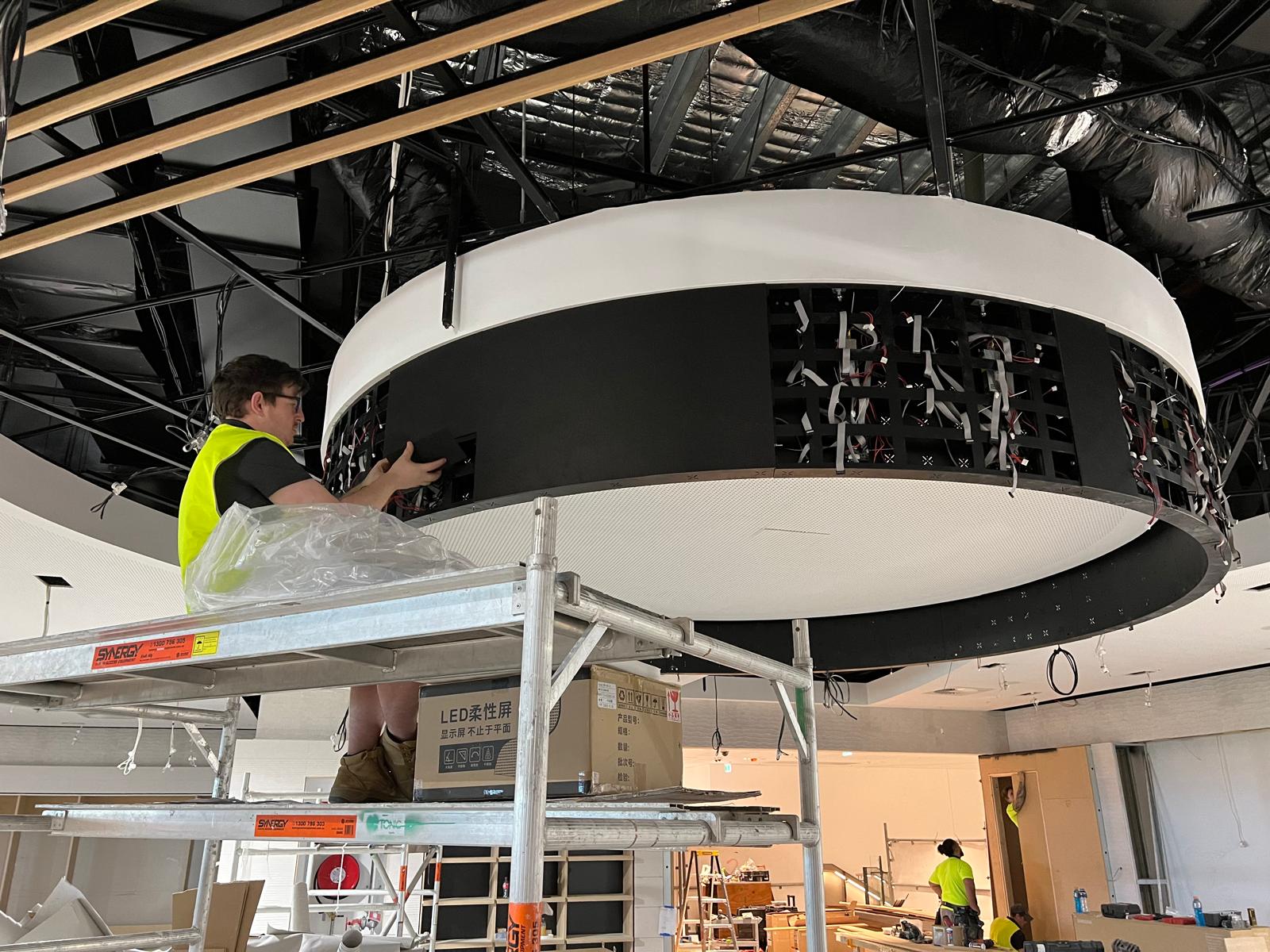An installer wiring the suspended halo LED ring from a scaffold platform, with the ceiling services still exposed overhead.
