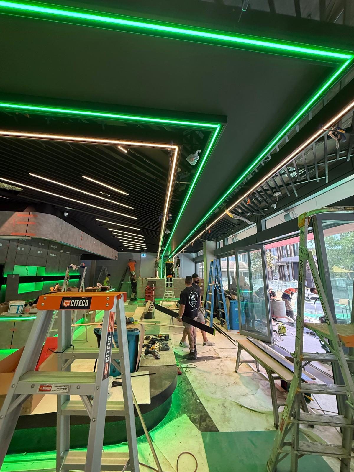 Triangular LED ribbon ceiling feature taking shape, with the strip lighting already glowing while the surrounding ceiling grid is still under construction.